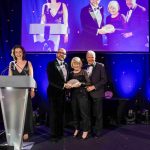 Presenter Gabriella Foley standing behind a lectern and co-presenter, Tim Rhys Evans presenting The Robert Maskrey Award for Arts Philanthropy to winners Alan and Sonja Jones